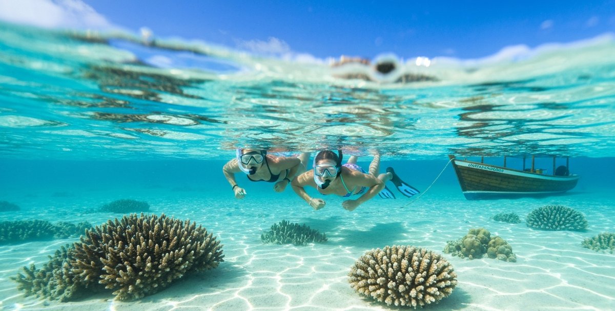 Visitors snorkelling in crystal-clear turquoise water near the sandbar islets of Île aux Cerfs, with white sand visible beneath the surface and a traditional wooden boat anchored nearby under bright midday sun