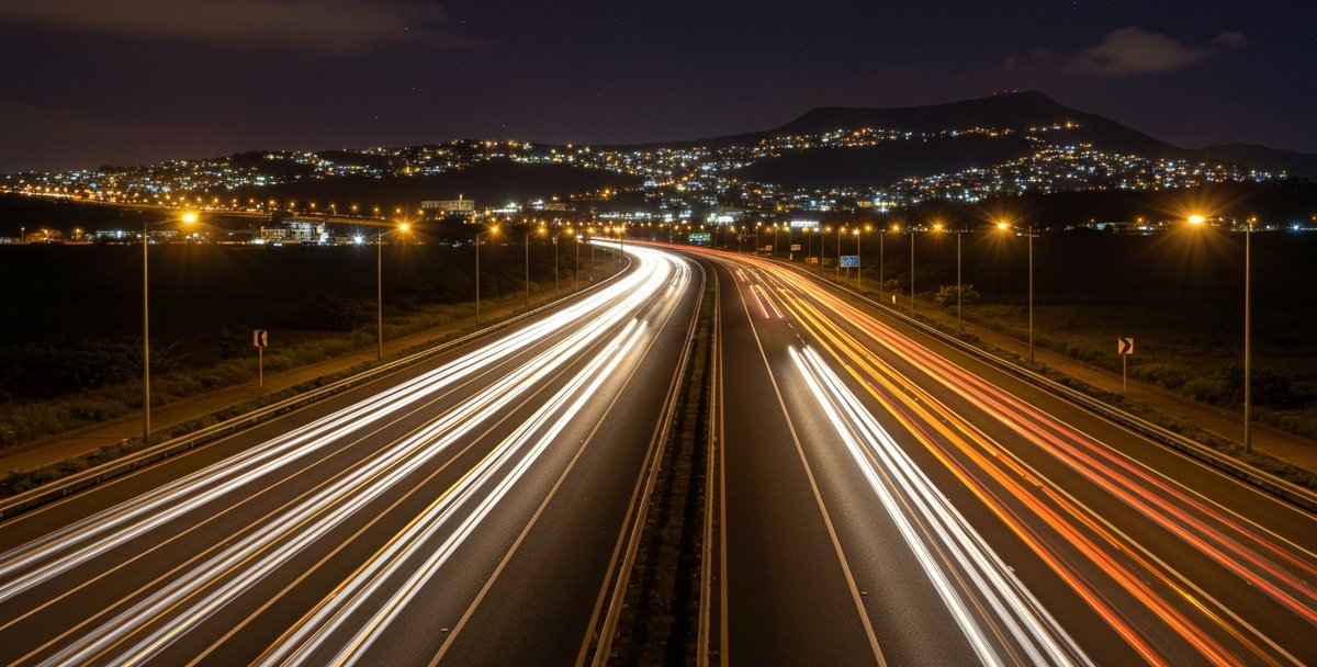 A well-lit stretch of the M1 motorway near Port Louis at night, with light trails from passing vehicles, the city's hillside lights visible in the distance, and a clear modern road surface
